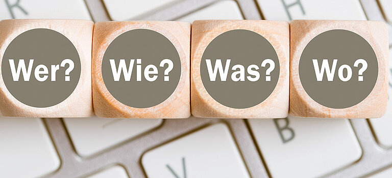 Das Bild zeigt vier Würfel, welche auf einer Tastatur liegen und die Wörter "Wer?", "Wie?", "Was?" und "Wo?" als Aufschrift haben.