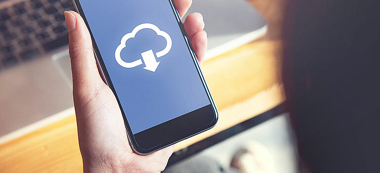 Das Bild zeigt ein Handy, welches in der Hand gehalten wird. Auf dem Handy sieht man eine Wolke und einen Pfeil nach unten (Download Symbol). 