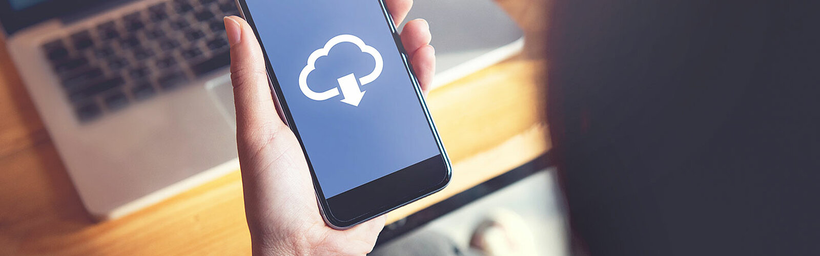 Das Bild zeigt ein Handy, welches in der Hand gehalten wird. Auf dem Handy sieht man eine Wolke und einen Pfeil nach unten (Download Symbol). 