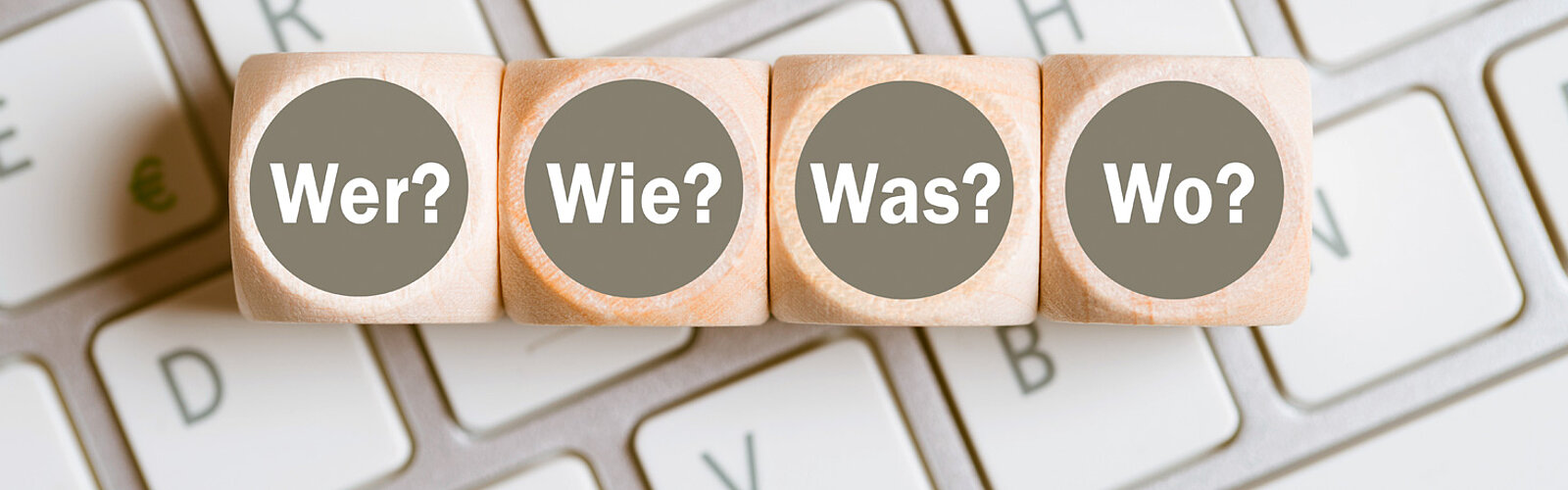 Das Bild zeigt vier Würfel, welche auf einer Tastatur liegen und die Wörter "Wer?", "Wie?", "Was?" und "Wo?" als Aufschrift haben.