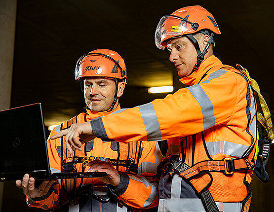 Das Bild zeigt zwei Arbeiter in Schutzausrüstung, die auf vor Ort auf einen Laptop schauen.