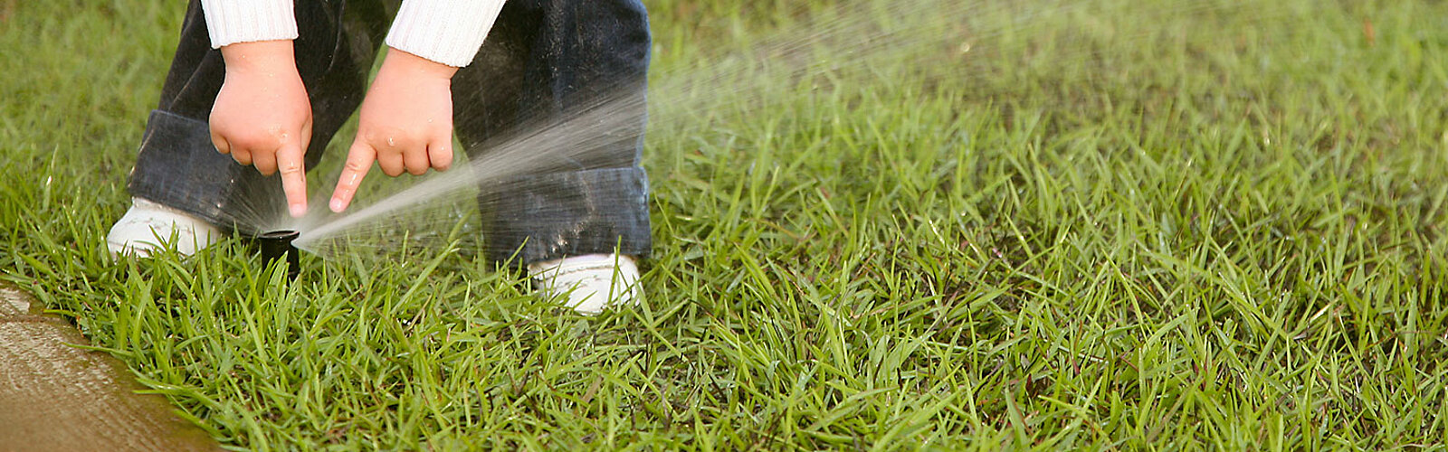 Dieses Bild zeigt eine Wiese mit einem Wassersprinkler, welcher von einem Kind angefasst wird.