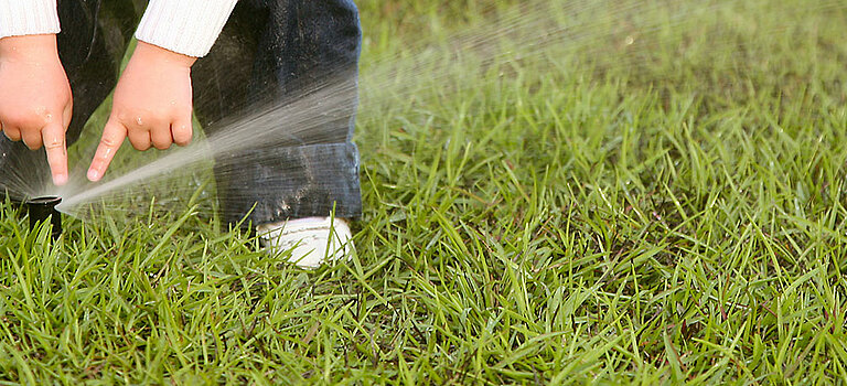 Dieses Bild zeigt eine Wiese mit einem Wassersprinkler, welcher von einem Kind angefasst wird.