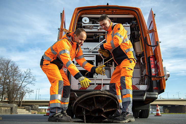 Dieses Bild zeigt zwei Mitarbeiter der Stadtentwässerung, die vor einem Fahrzeug einen Schachtdeckel anheben.