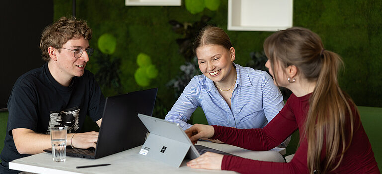 Studierende und Trainees tauschen sich an einem Tisch aus und arbeiten gemeinsam mit Laptop und Tablet.