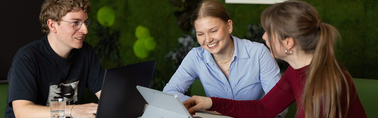 Studierende und Trainees tauschen sich an einem Tisch aus und arbeiten gemeinsam mit Laptop und Tablet.